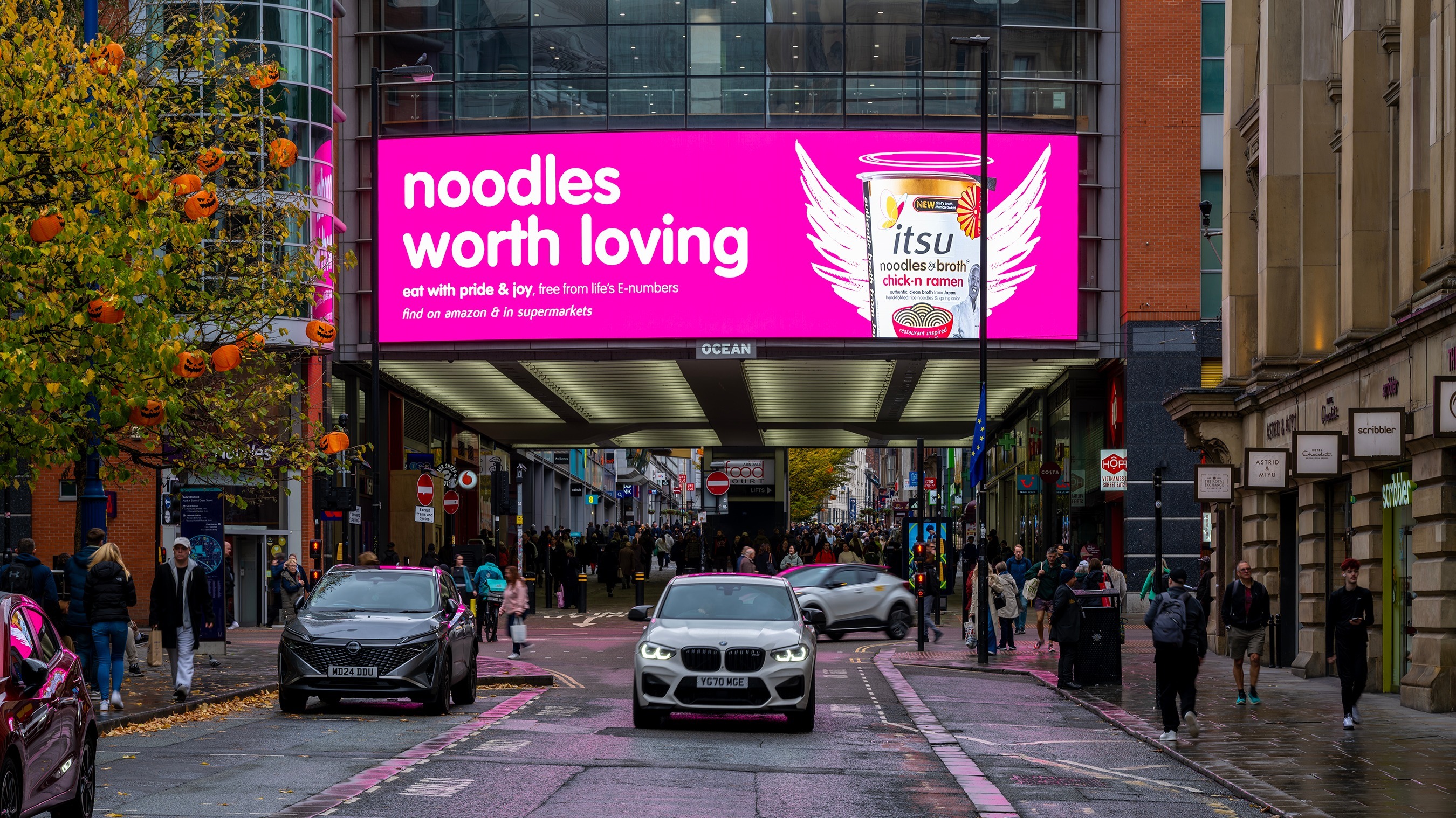 Pink billboard advertising 'noodles worth loving' overlooks a bustling city street with vehicles.