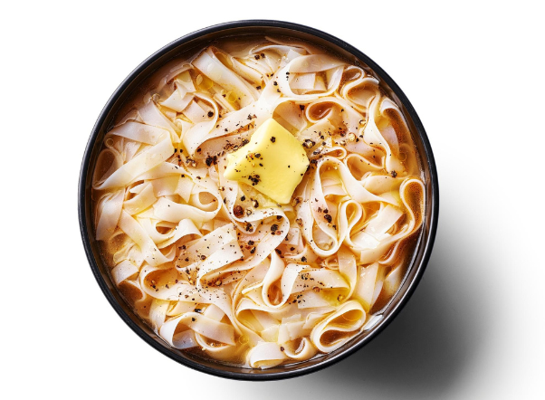 Overhead view of wide flat noodles in broth with melting butter and black pepper.