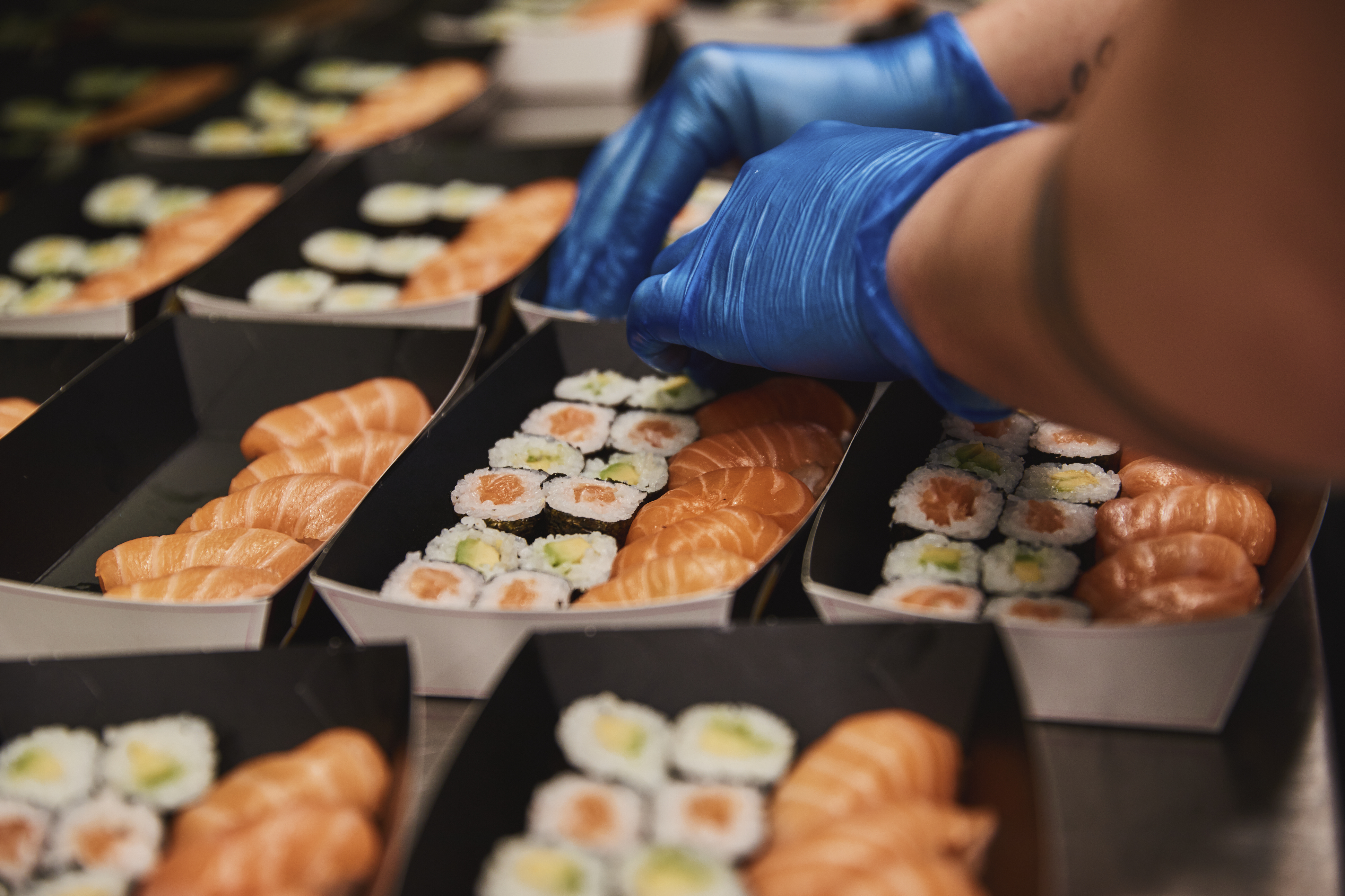 A gloved hand arranges salmon nigiri and maki sushi rolls into multiple catering trays.