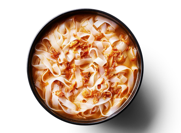 A close-up of wide flat noodles in an orange broth, topped with crispy fried shallots.