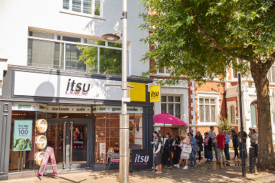 A long queue of people waits patiently outside an itsu restaurant on a bright, sunny day.