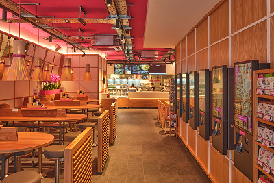 Modern restaurant interior featuring wooden tables, pink ceiling, and a service counter in the background.
