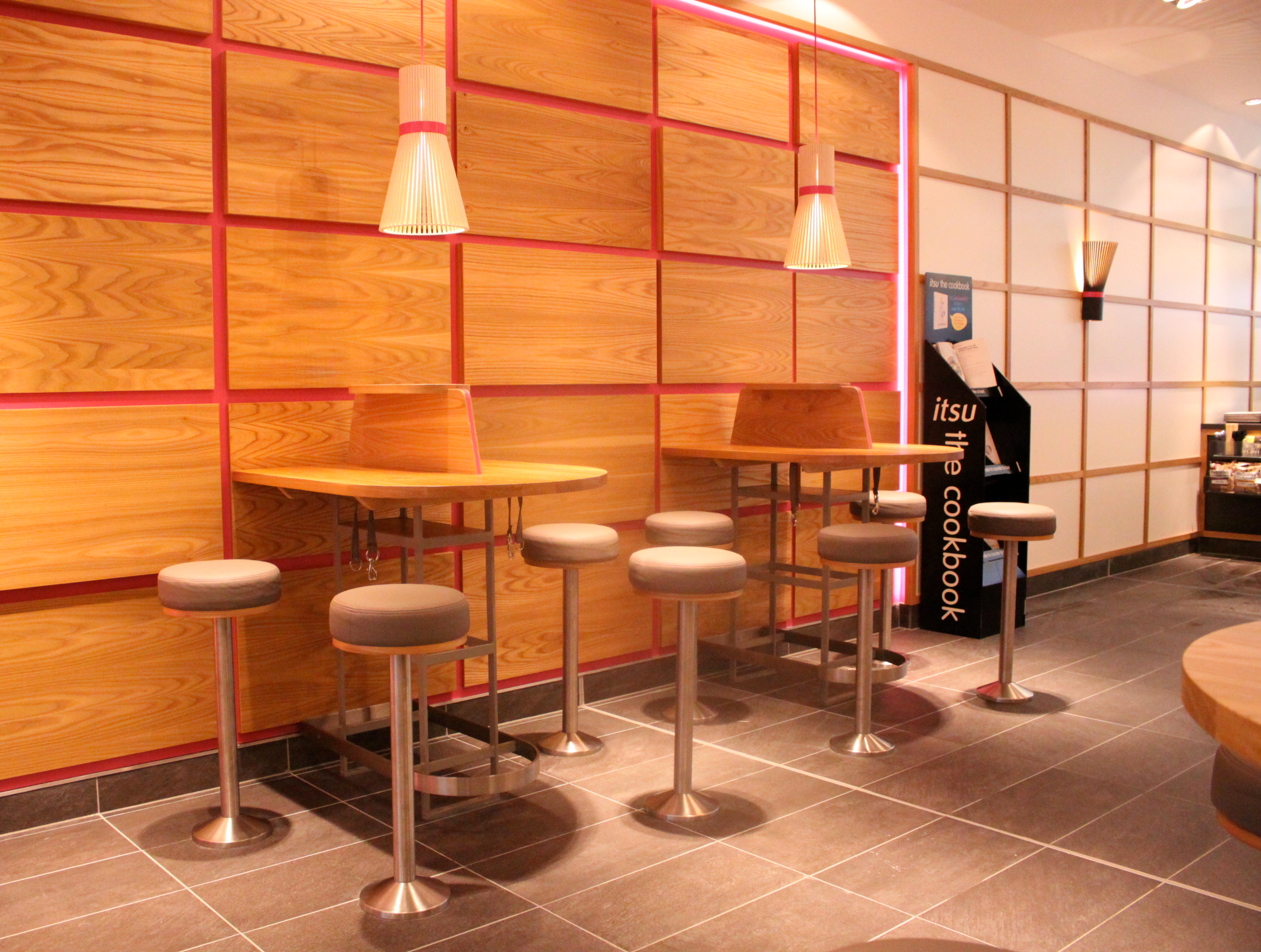 Modern interior with high wooden tables, grey stools, and light wood panelled walls.