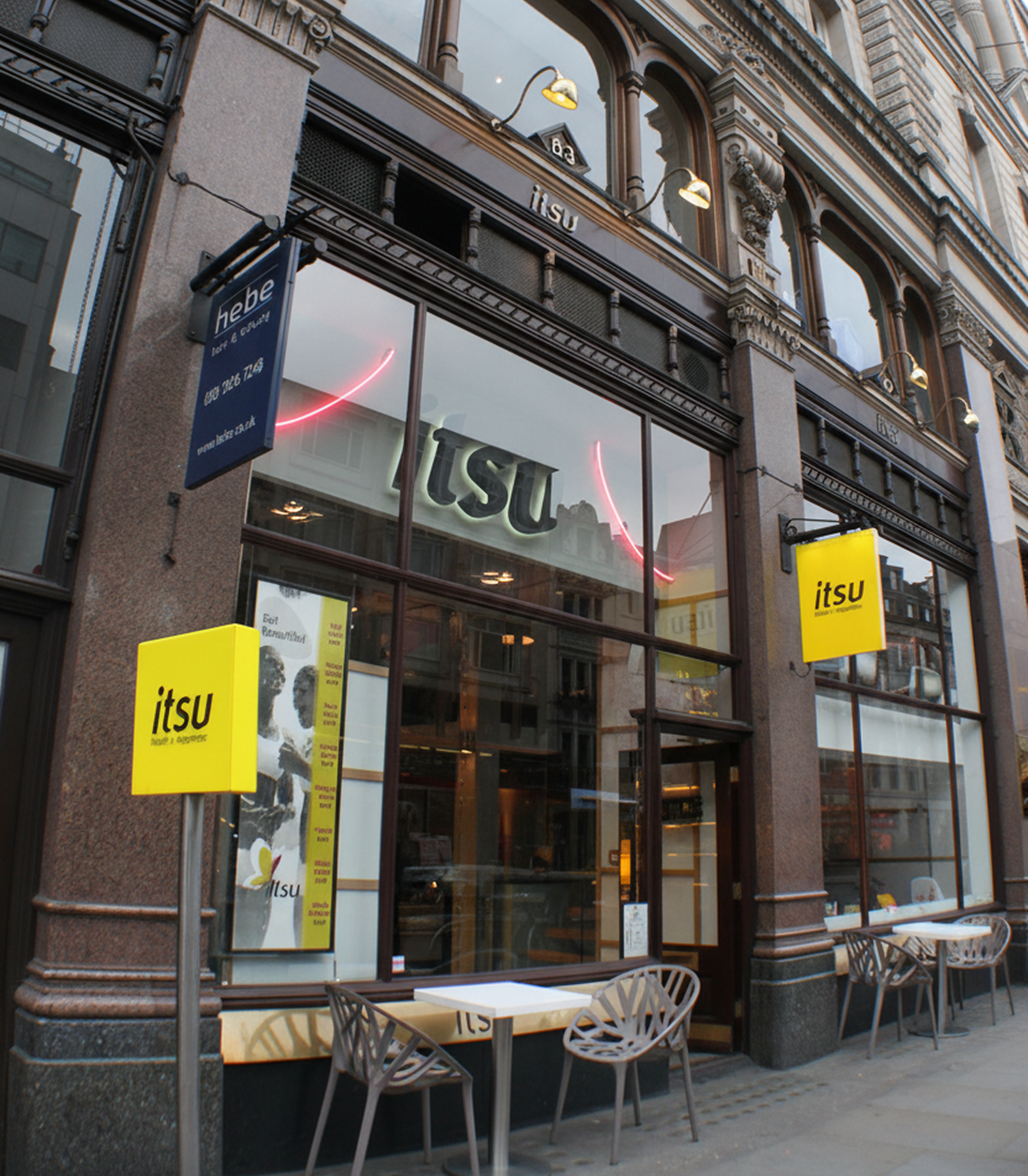 Modern Itsu restaurant exterior with outdoor tables and yellow signs on a city street.