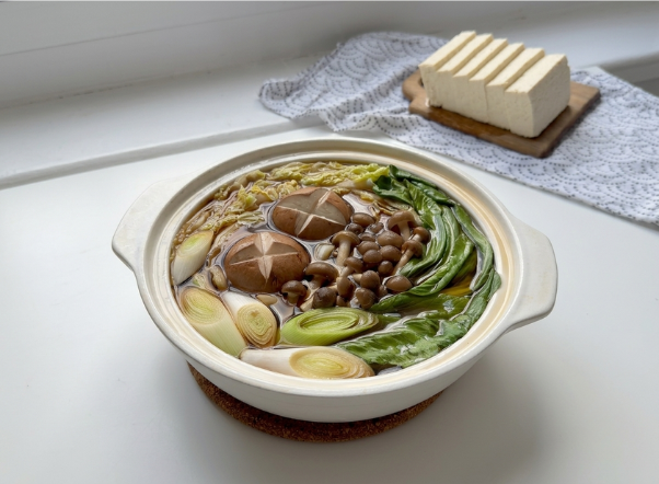 Japanese hot pot with shiitake mushrooms, leafy greens, leeks, and smaller mushrooms, with tofu block nearby.