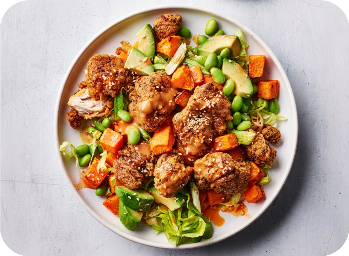 A vibrant bowl features crispy fried chicken, sweet potatoes, edamame, and avocado slices.