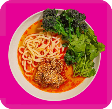 A bowl of udon noodles with broccoli, cilantro, and sesame-crusted protein in red broth.