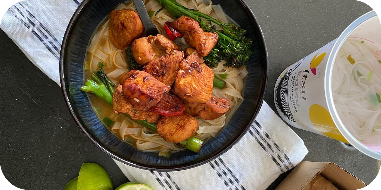 Noodles with chicken, broccoli, and peppers in a bowl next to instant noodle cup.