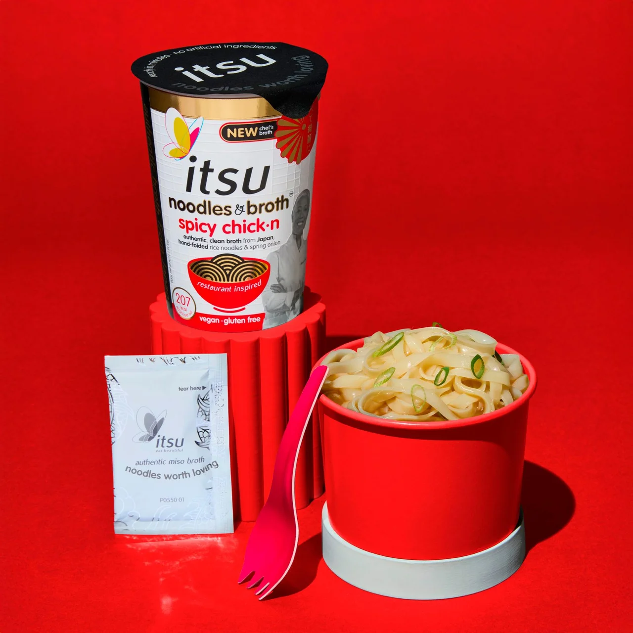 Itsu spicy chicken noodle cup, cooked noodles, seasoning packet, and pink fork.