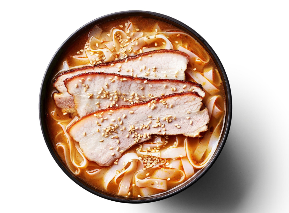 Dark bowl of ramen featuring chashu pork slices, wide flat noodles, and sesame seeds.
