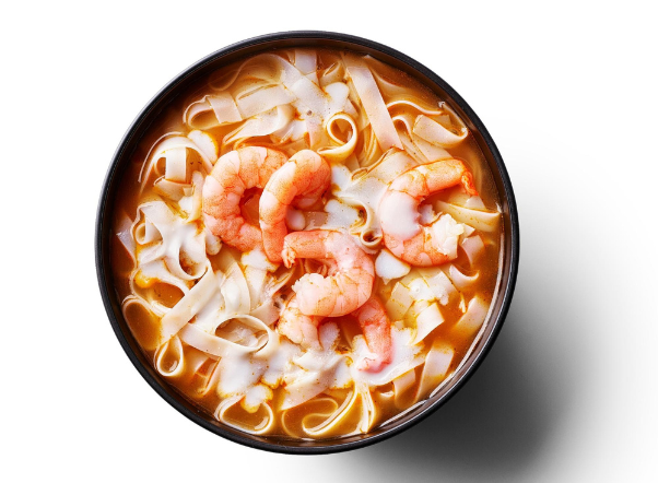 A black bowl of shrimp noodle soup with flat rice noodles and a reddish broth.