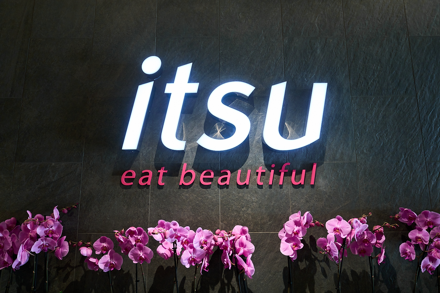 Glowing 'itsu eat beautiful' sign above vibrant purple orchids on a dark wall.