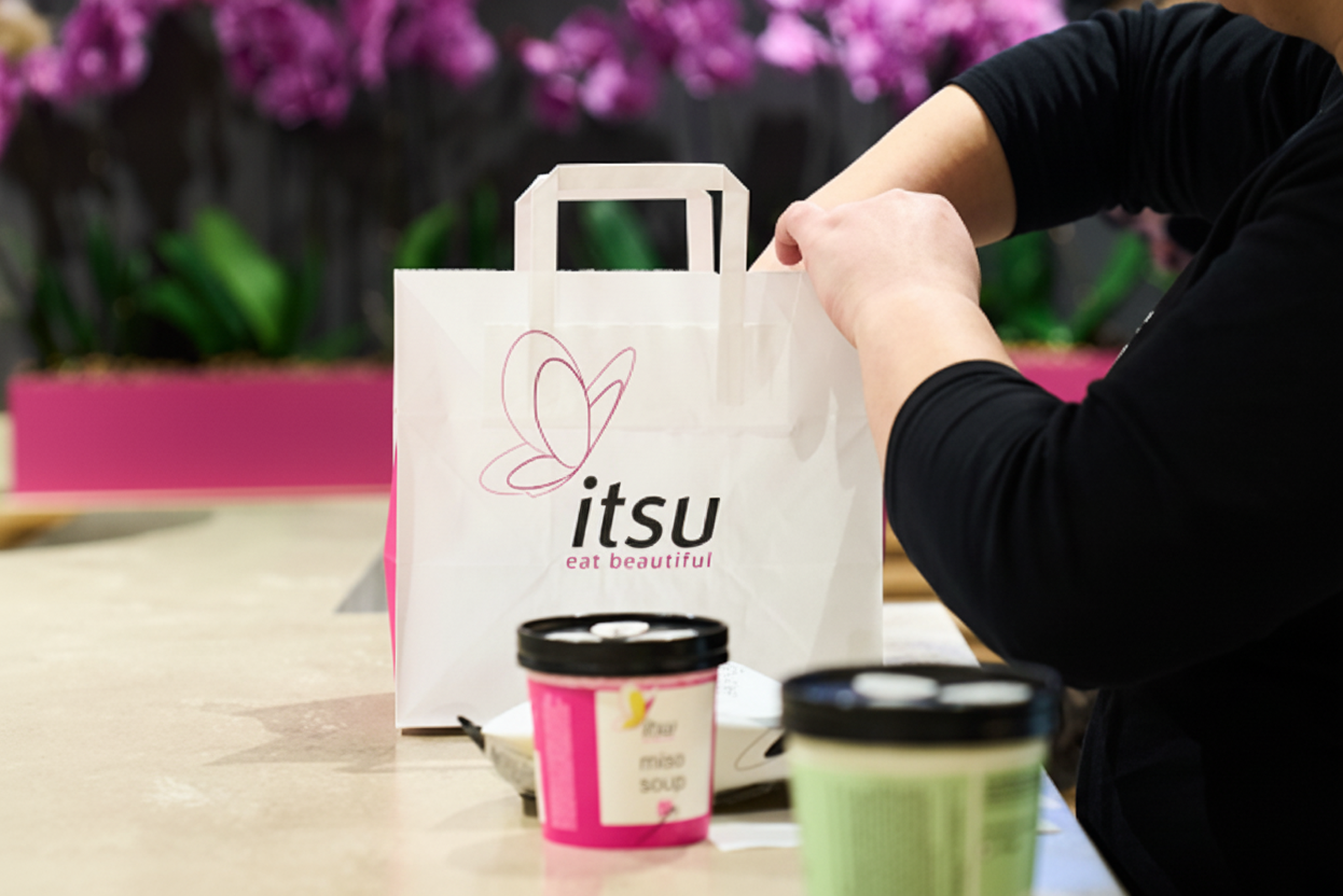 Person placing items into an Itsu branded paper bag with food containers on a counter.