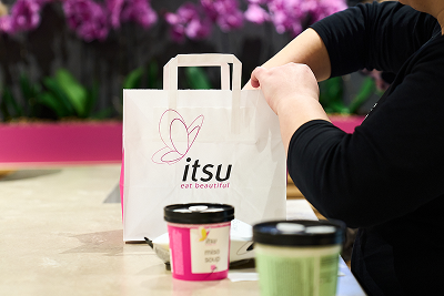 Person placing items into an Itsu paper bag with miso soup containers on a table.