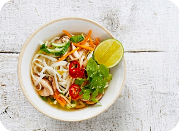 A bowl of pho soup with noodles, vegetables, mushrooms, and a lime wedge.
