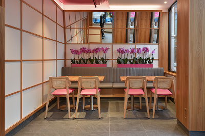 Modern restaurant interior featuring wooden tables, pink-seated chairs, and floral decorations under grid-paneled walls.