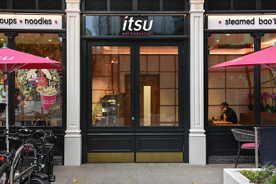 itsu restaurant storefront with black doors, pink umbrellas, and a person sitting inside.