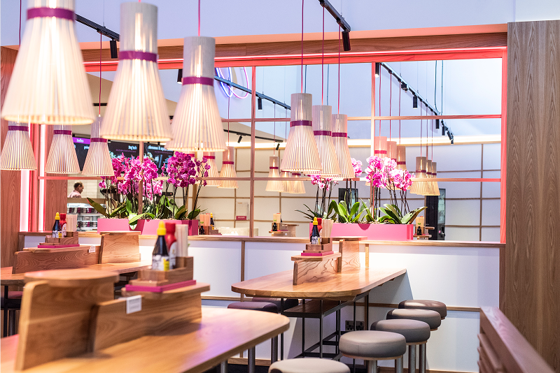 Modern restaurant interior with wooden tables, gray stools, and vibrant pink orchid decorations.