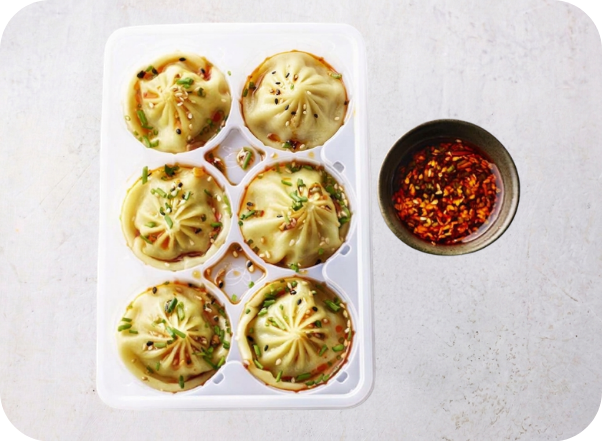 Six soup dumplings garnished with green onions and chili flakes, served with a side of chili oil.