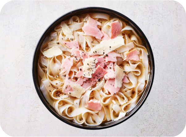 A close-up of flat noodles in broth topped with ham, cheese, and black pepper.