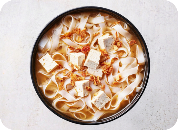 Top-down view of a bowl of noodle soup with tofu, flat noodles, and crispy toppings.