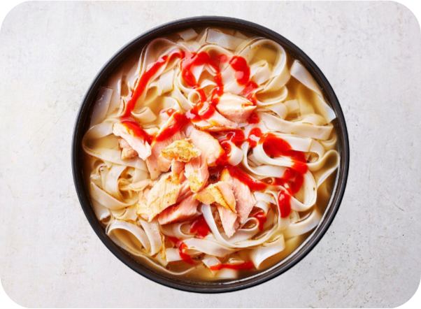 Bowl of pho noodle soup with salmon, wide rice noodles, and a spicy red sauce.