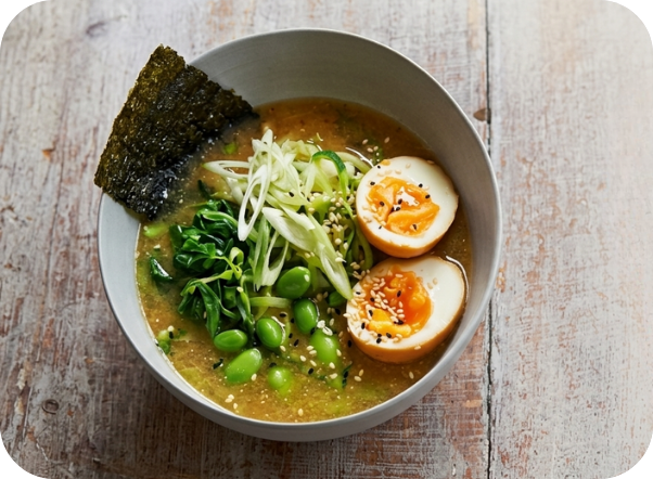 Bowl of ramen with soft-boiled eggs, edamame, spinach, and nori on a wooden table.