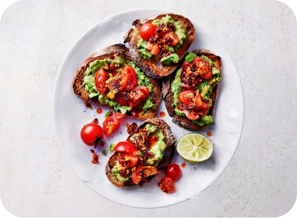 Avocado toast with diced tomatoes, spices, and a lime wedge on a white plate.