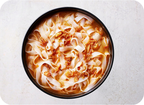 A bowl of wide rice noodles in a savory broth, topped with fried shallots.