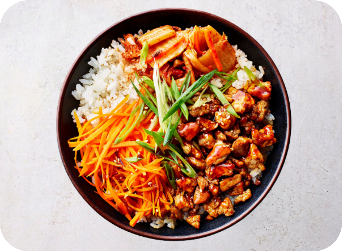 Vibrant bowl with white rice, kimchi, shredded carrots, green onions, and glazed chicken.