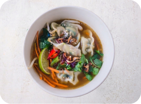 A vibrant bowl of dumpling soup garnished with lime, chili, carrots, and fresh herbs.