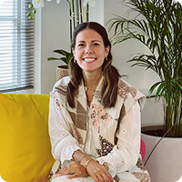 Smiling woman in a patterned top, seated indoors with a yellow cushion and plants.