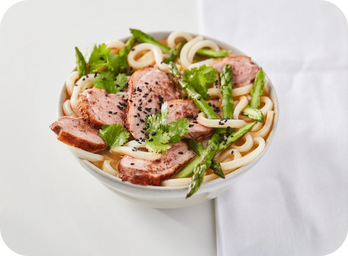 Bowl of udon noodles with sliced duck, asparagus, cilantro, and black sesame seeds.