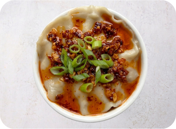 Pork dumplings in a white bowl, topped with spicy chili oil and chopped green onions.