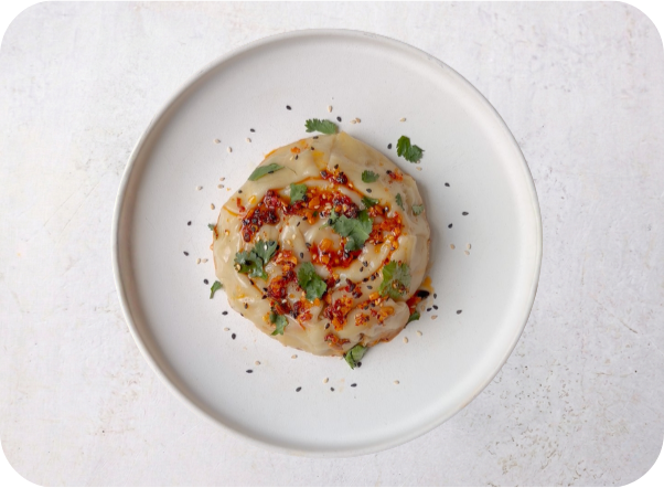 Round savory pastry topped with chili oil, cilantro, and seeds on a white plate.