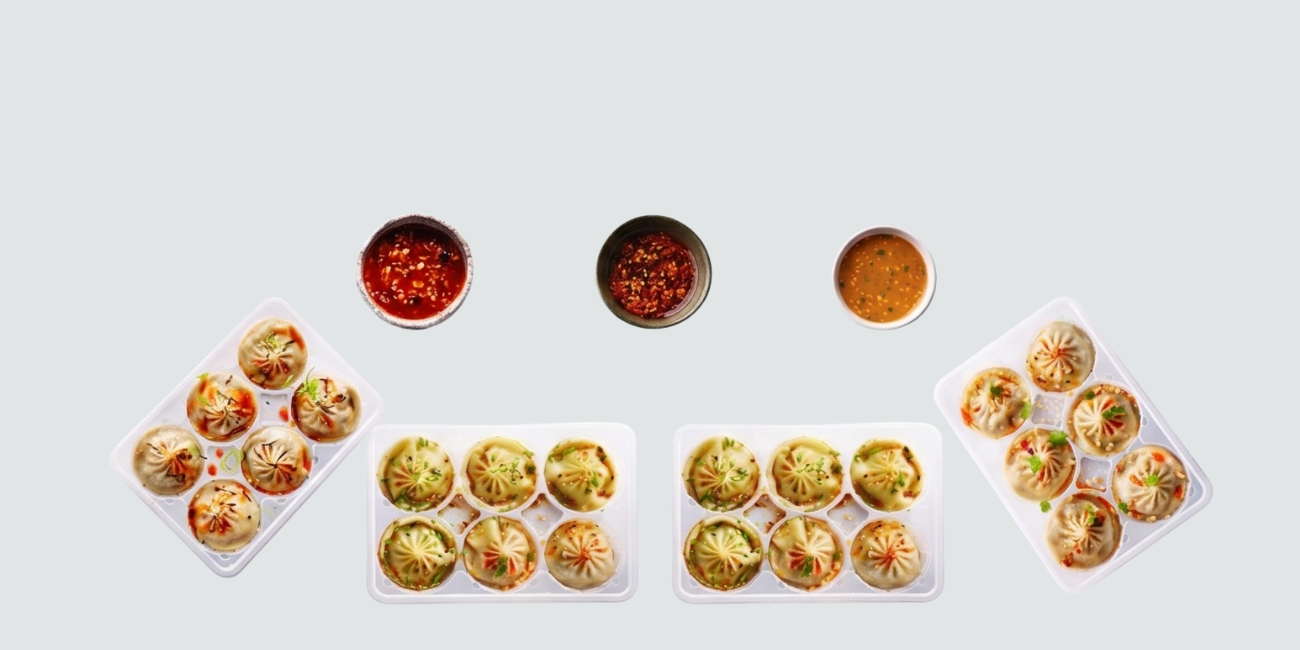 Assortment of savory dumplings in trays with three dipping sauces on a gray background.
