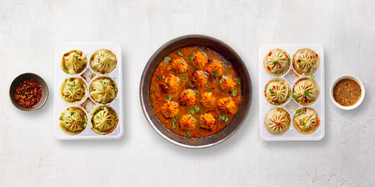 Kofta curry in a bowl, surrounded by two trays of momos and dipping sauces.
