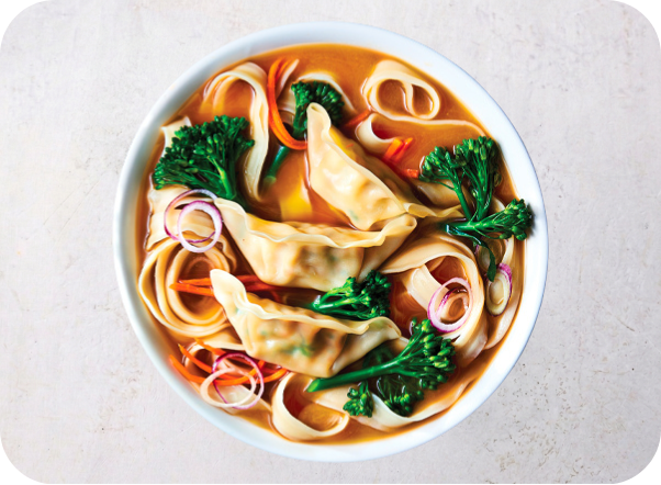 A bowl of savory dumpling soup with noodles, broccoli, shredded carrots, and red onion.