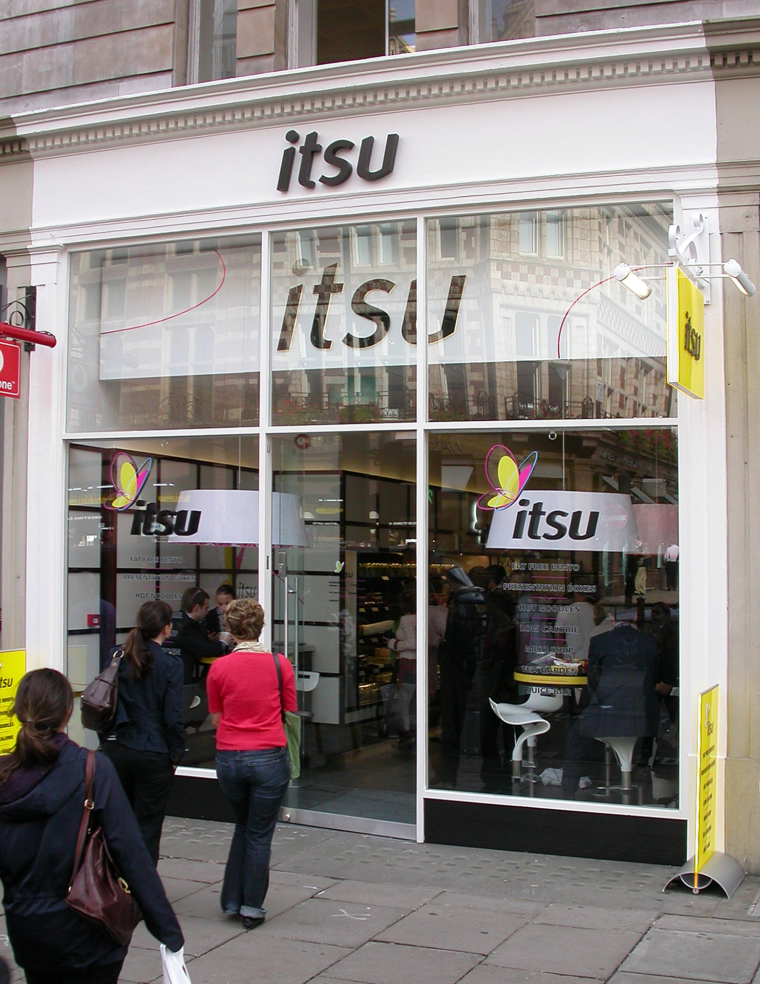 Itsu restaurant storefront with a white facade and large windows, people entering the entrance.