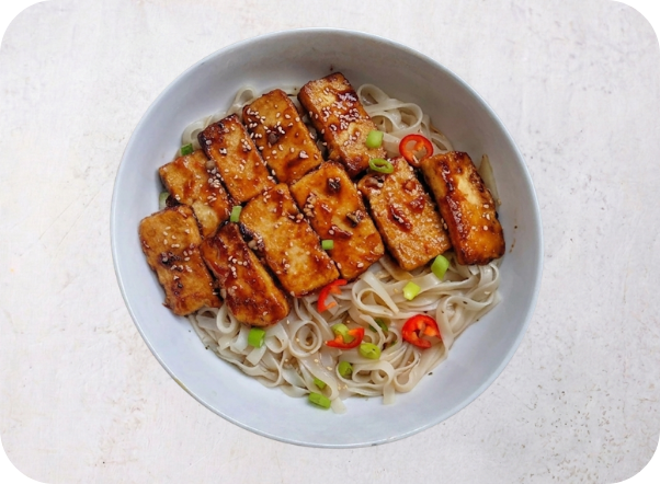 Pan-fried tofu slices glazed with sauce, garnished with green onions and chili, served over noodles.