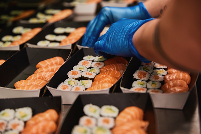 Gloved hands prepare trays of fresh salmon nigiri and sushi rolls for packaging.