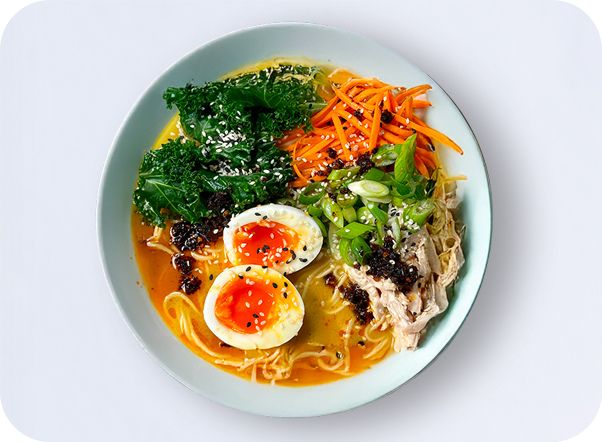 Vibrant ramen bowl with noodles, rich broth, kale, carrots, chicken, and soft-boiled eggs.