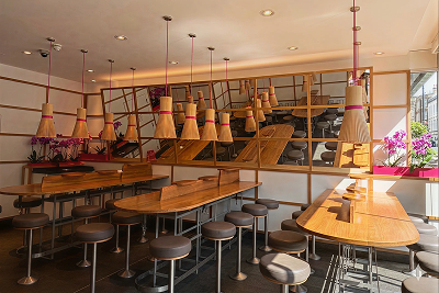 Modern restaurant interior with wooden tables, brown stools, unique bamboo lights, and pink orchid decorations.