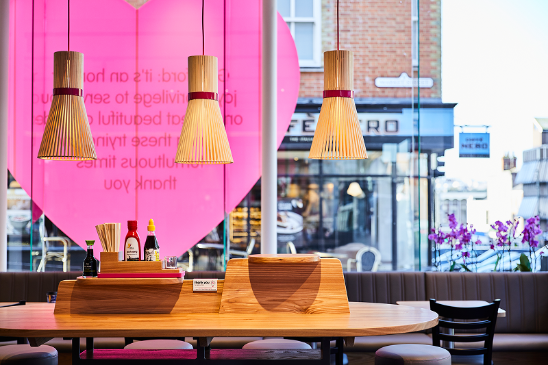 Restaurant interior with wooden table, unique pendant lights, and a large pink heart window.