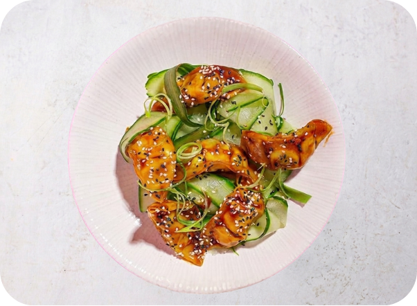 Glossy teriyaki chicken with sesame seeds atop fresh cucumber ribbons in a pink bowl.