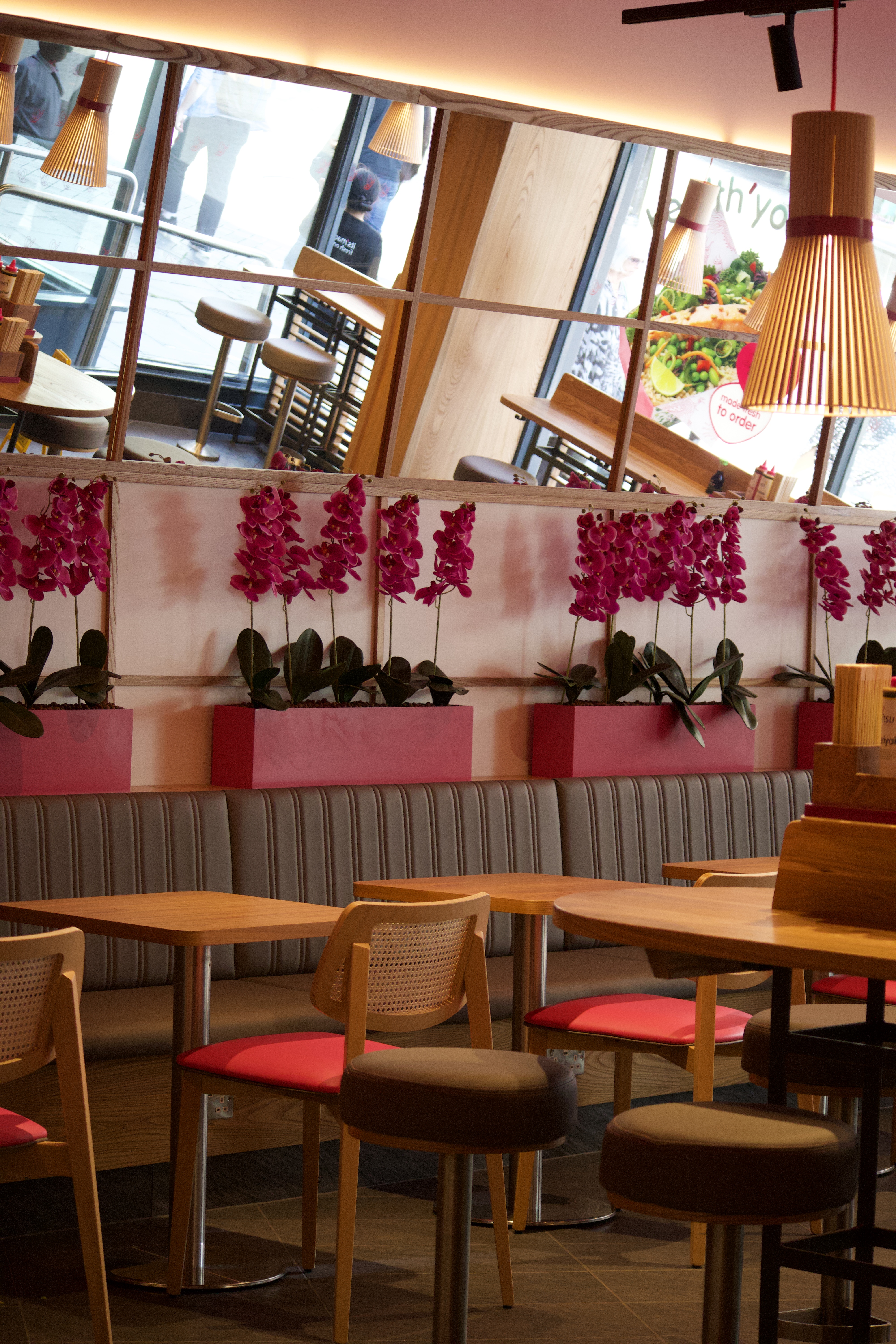 Modern restaurant interior with wooden tables, chairs, pink decorative wall, and large reflective windows.