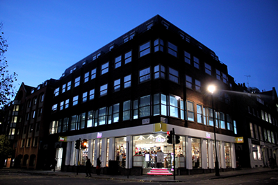 Modern building at dusk with illuminated retail storefront on ground floor and dark sky.