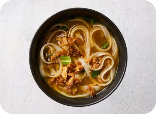Bowl of savory noodle soup with flat noodles, crispy fried shallots, and green onions.