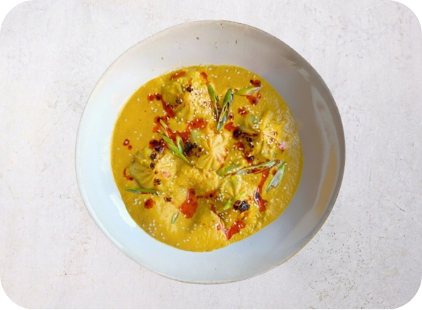Yellow creamy curry with dumplings, red oil, and green scallions in a white bowl.