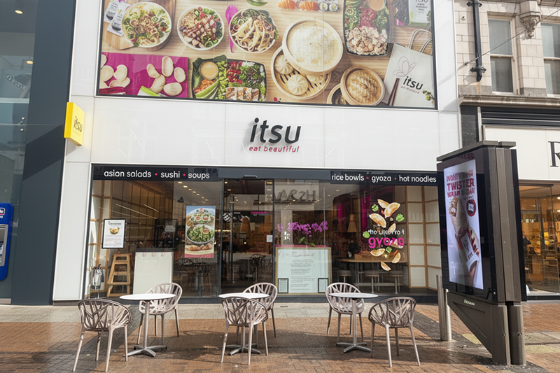 Itsu restaurant exterior with outdoor seating and a large food display above.